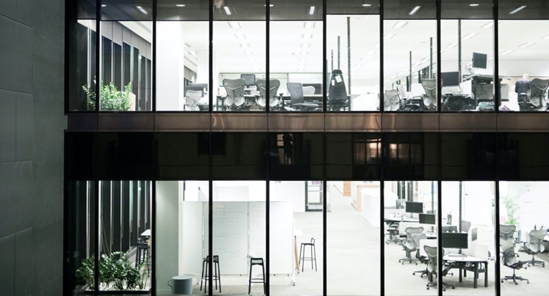 Exterior of the windows looking into work chairs and desks on two levels of an empty interior commercial office at night